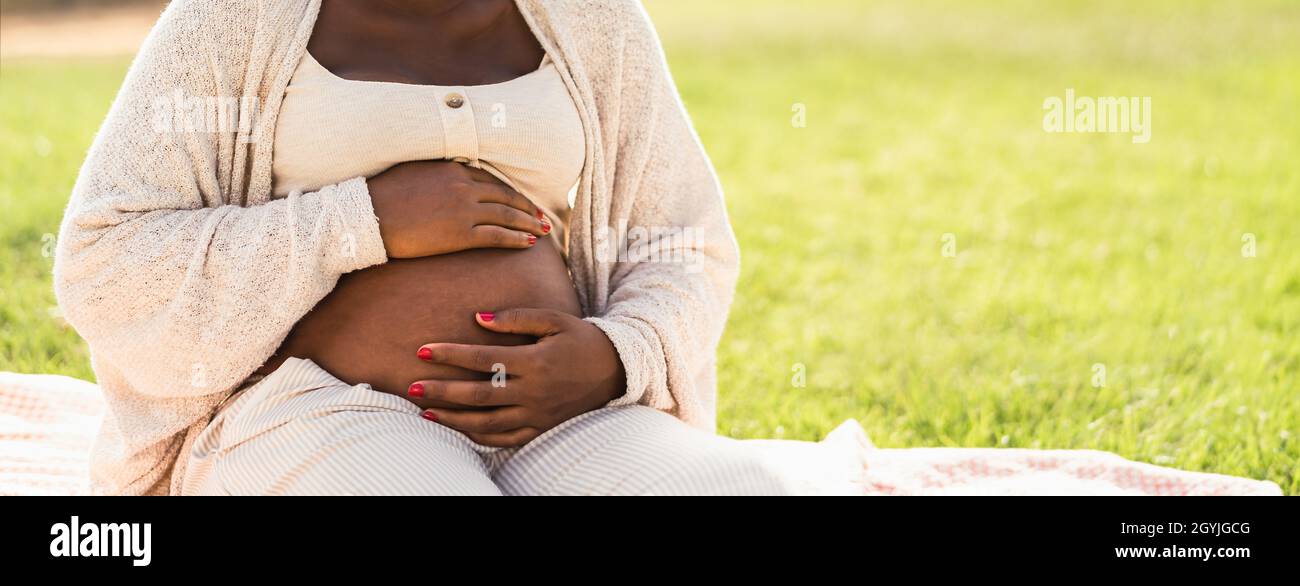 Primo piano su ventre incinta di giovane donna africana seduta nel parco - concetto di stile di vita di maternità Foto Stock
