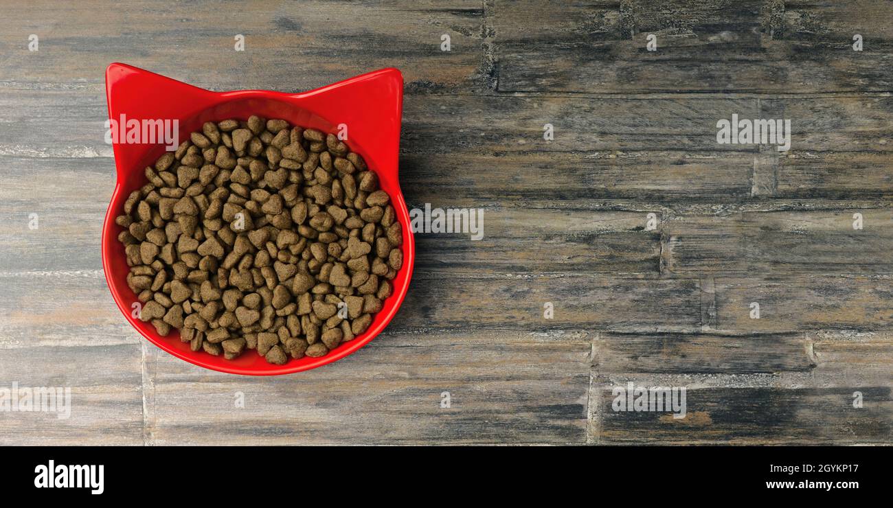 Ciotola rossa a forma di gatto riempita con cibo secco per animali domestici su sfondo rustico. Immagine panoramica con spazio di copia. Foto Stock