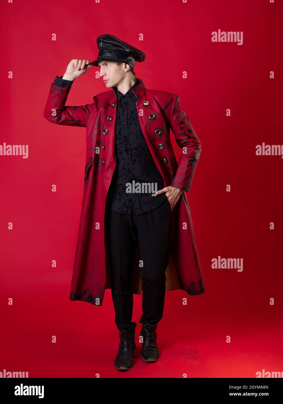 Un giovane oltraggioso in un audace cappotto rosso e in un berretto in pelle nera, stile militare, in stile nero, foto su sfondo rosso Foto Stock