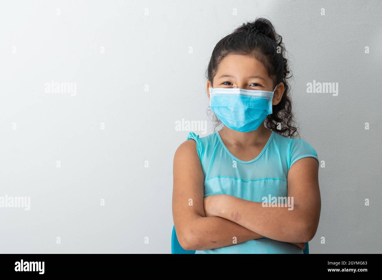 bambina (8 anni) guardando verso la macchina fotografica sorridente, aspetto felice, seduta con le braccia incrociate e maschera chirurgica blu. Medico, farmaceutico e Foto Stock