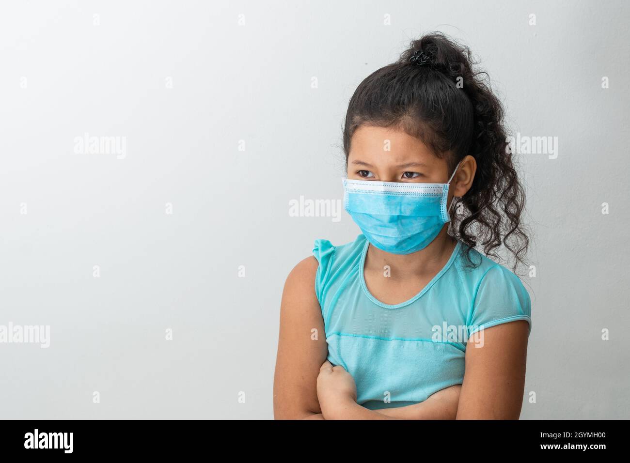 bambina (8 anni) seduta con le braccia incrociate, ragazza marrone arrabbiata con una maschera chirurgica blu. Concetto medico, farmaceutico e sanitario. Foto Stock