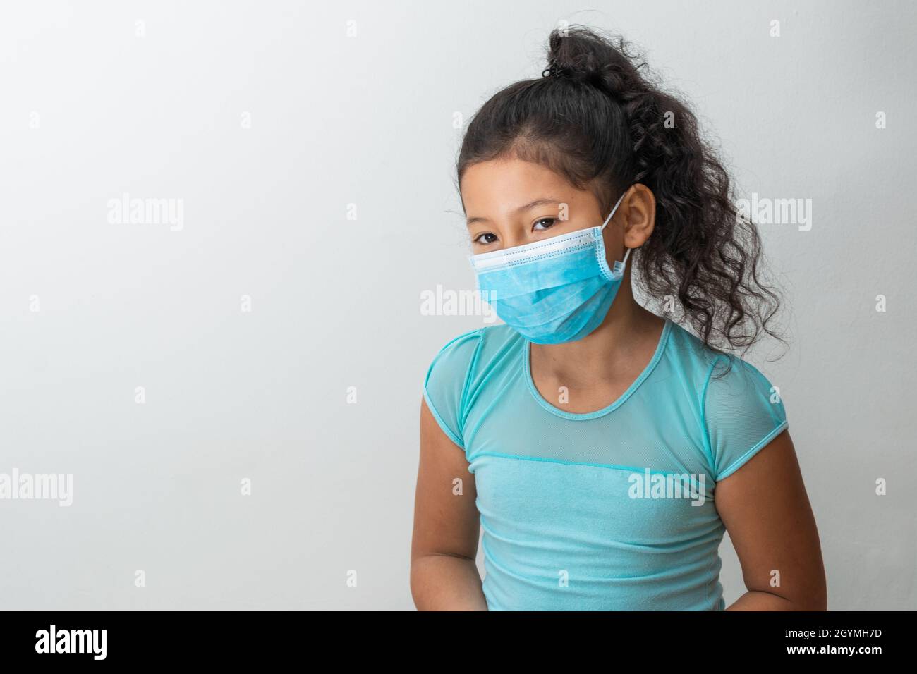 bambina (8 anni) seduta con le braccia giù marrone ragazza pensiva ricevere una notizia con una maschera chirurgica blu. Medico, farmaceutico e sanita Foto Stock