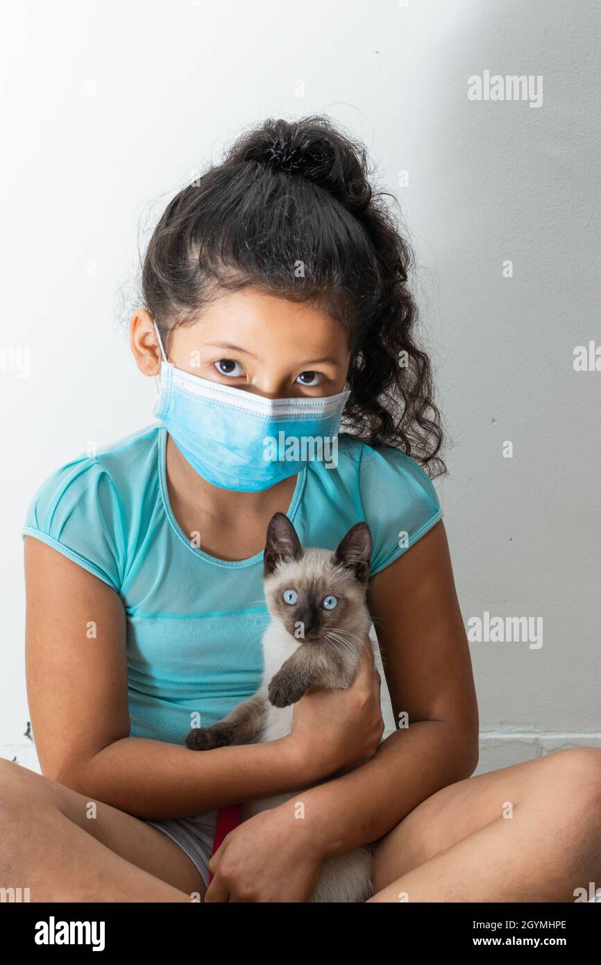 Bambina (8 anni) seduta con un gatto siamese nelle mani, ragazza marrone con una maschera chirurgica blu. Concetto medico, farmaceutico e sanitario. Foto Stock