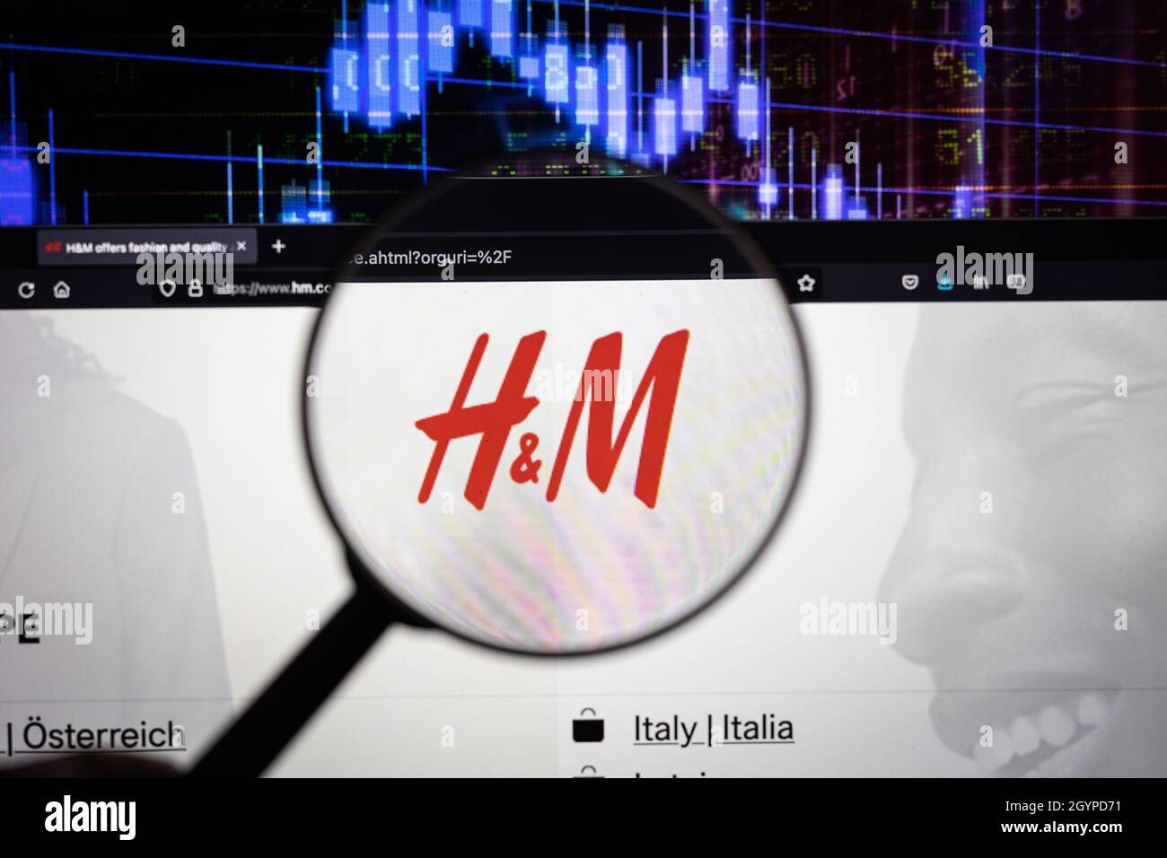 Logo dell'azienda H&M su un sito web con sviluppi del mercato azionario sfocati in background, visto su uno schermo del computer attraverso una lente d'ingrandimento Foto Stock