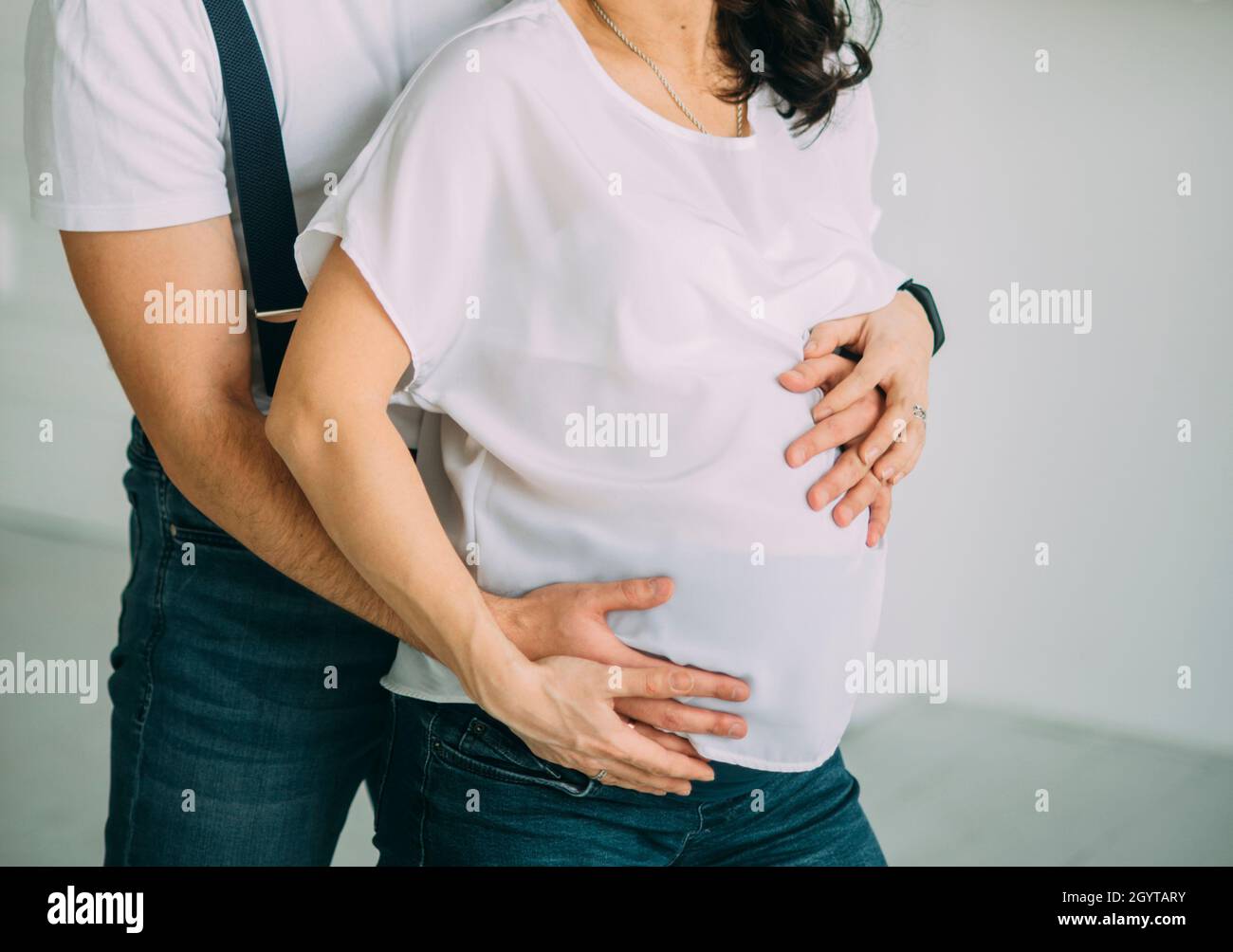 la coppia incinta abbraccia il ventre con le mani in camicia bianca e jeans blu capelli scuri Foto Stock