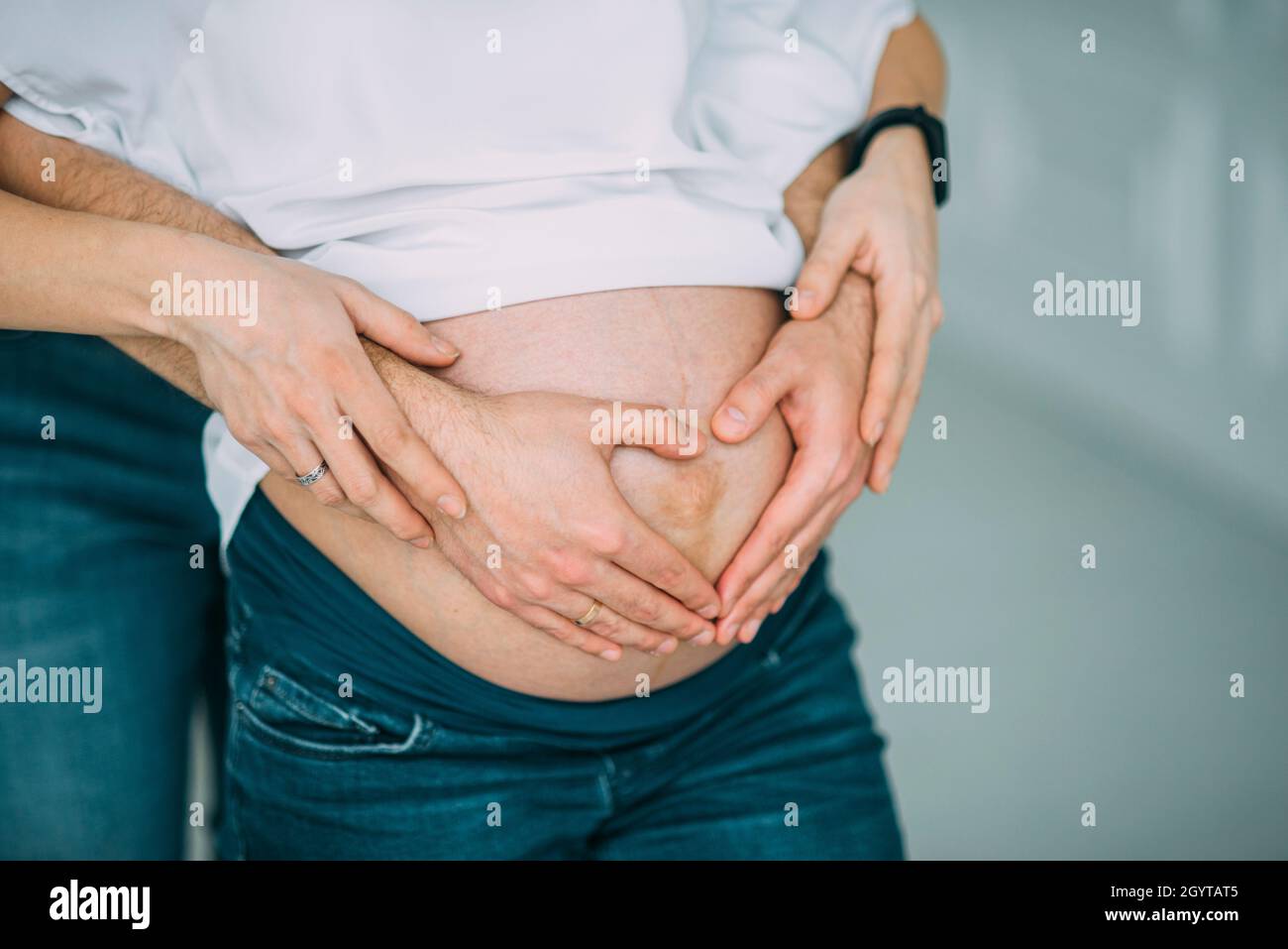 la coppia incinta abbraccia il ventre con le mani in camicia bianca e jeans blu anelli cuore Foto Stock