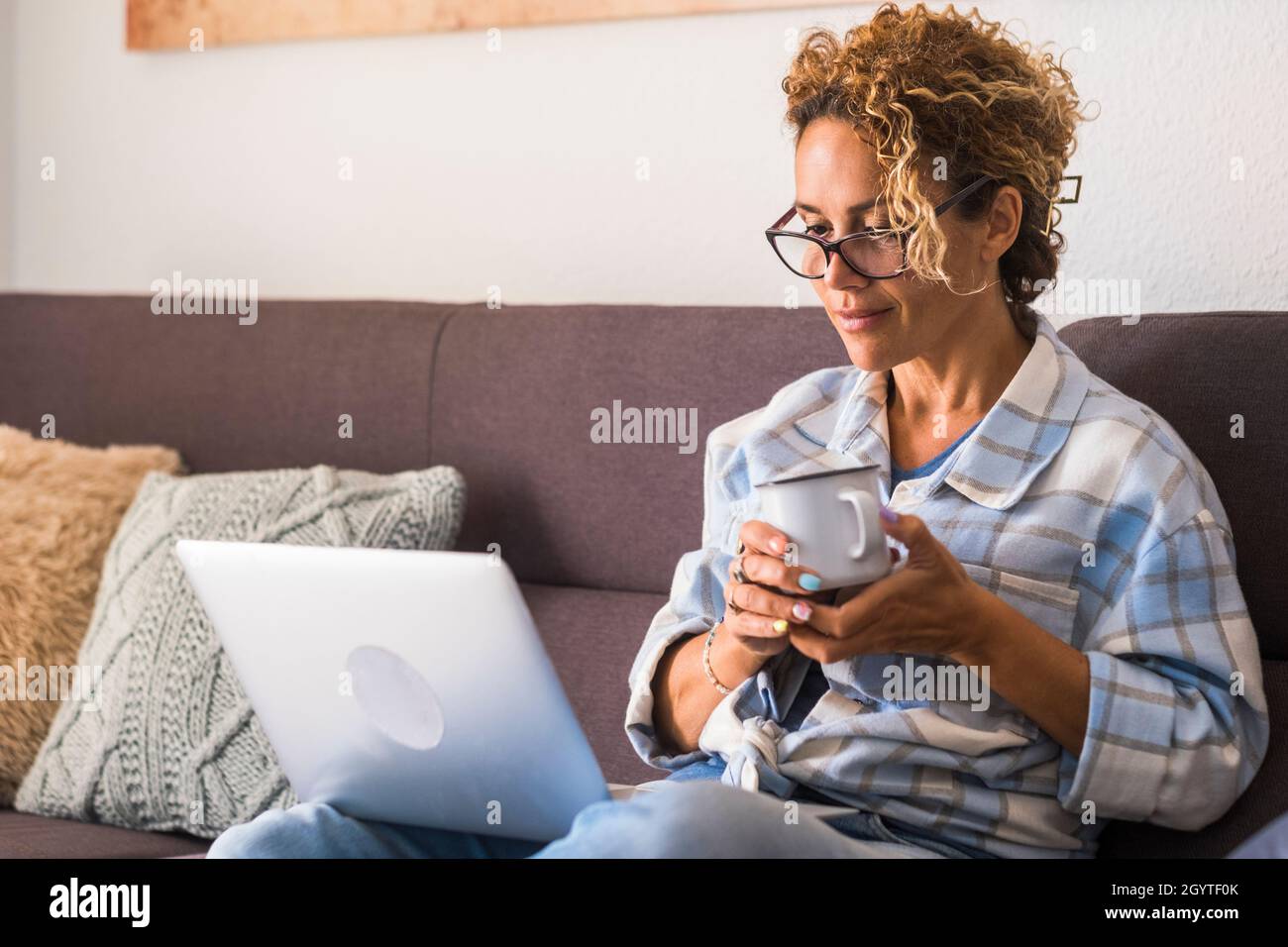 Bella giovane donna in occhiali che lavora su un computer portatile mentre beve il caffè seduto sul divano a casa. Donna d'affari in casuals e occhiali che lavorano f Foto Stock