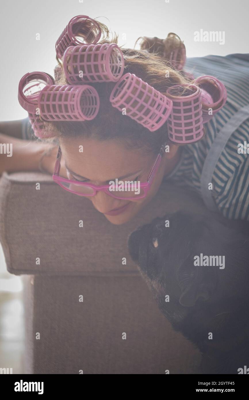 Giovane donna con curlers sui capelli e giocando con il suo cane da compagnia appoggiato sul divano in salotto a casa. Bella donna in occhiali spendendo tempo libero t Foto Stock