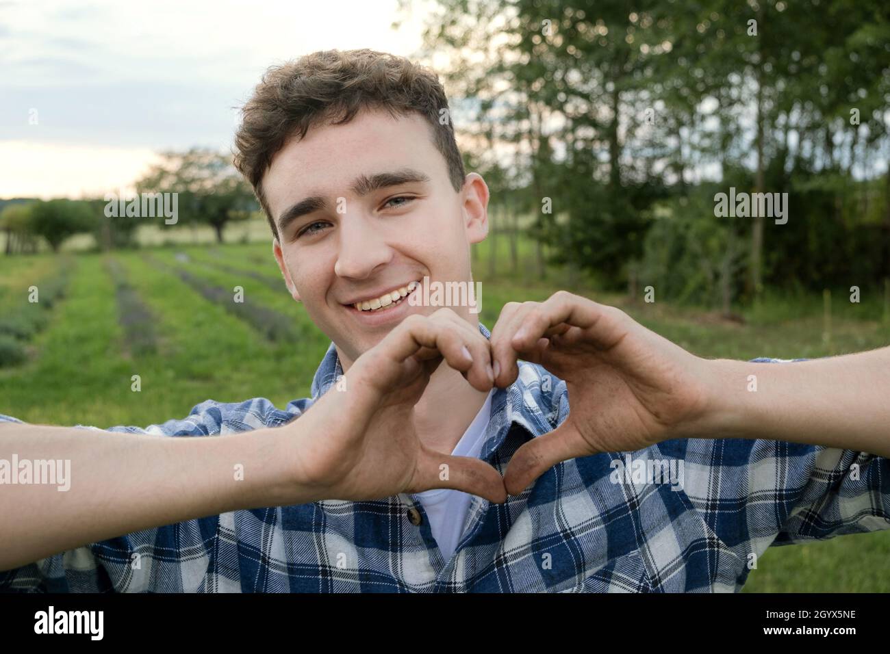 Giovane agricoltore sorridente mostra un gesto di forma cardiaca con amore per l'agricoltura. Buon lavoratore agricolo positivo con mani sporche. Foto Stock