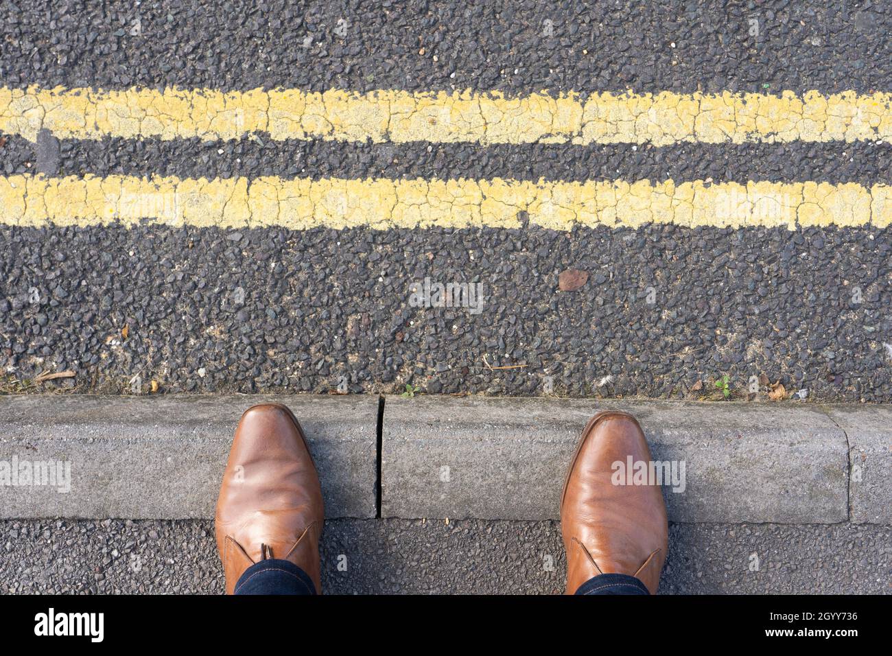 Piedi da uomo con scarpe in piedi al bordo di un marciapiede e marciapiede, accanto a una strada principale con due linee gialle. REGNO UNITO. Concetto - in attesa, sul bordo Foto Stock