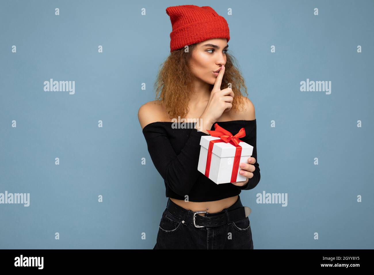 Affascinante felice adulto scuro biondo donna riccia isolato su blu parete  di fondo con elegante crop top nero e cappello rosso tenendo la scatola del  regalo che guarda a Foto stock -