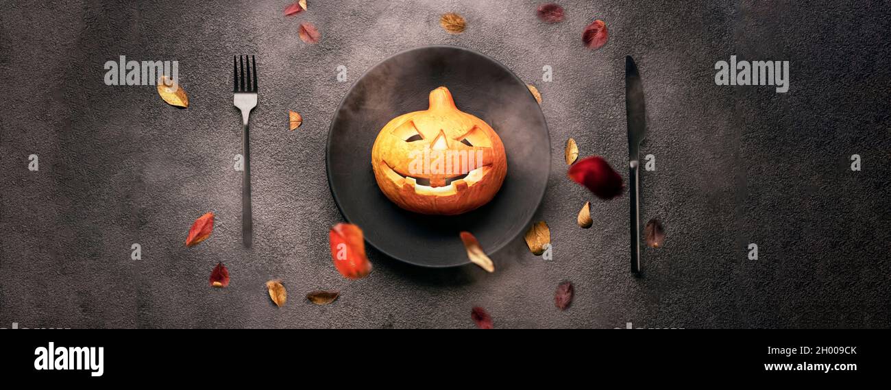 Zucca con occhi ardenti per Halloween su un piatto nero con forchetta e cucchiaio. Caduta foglie autunnali. Vacanze spaventose, orrore, foto panoramica. Foto Stock