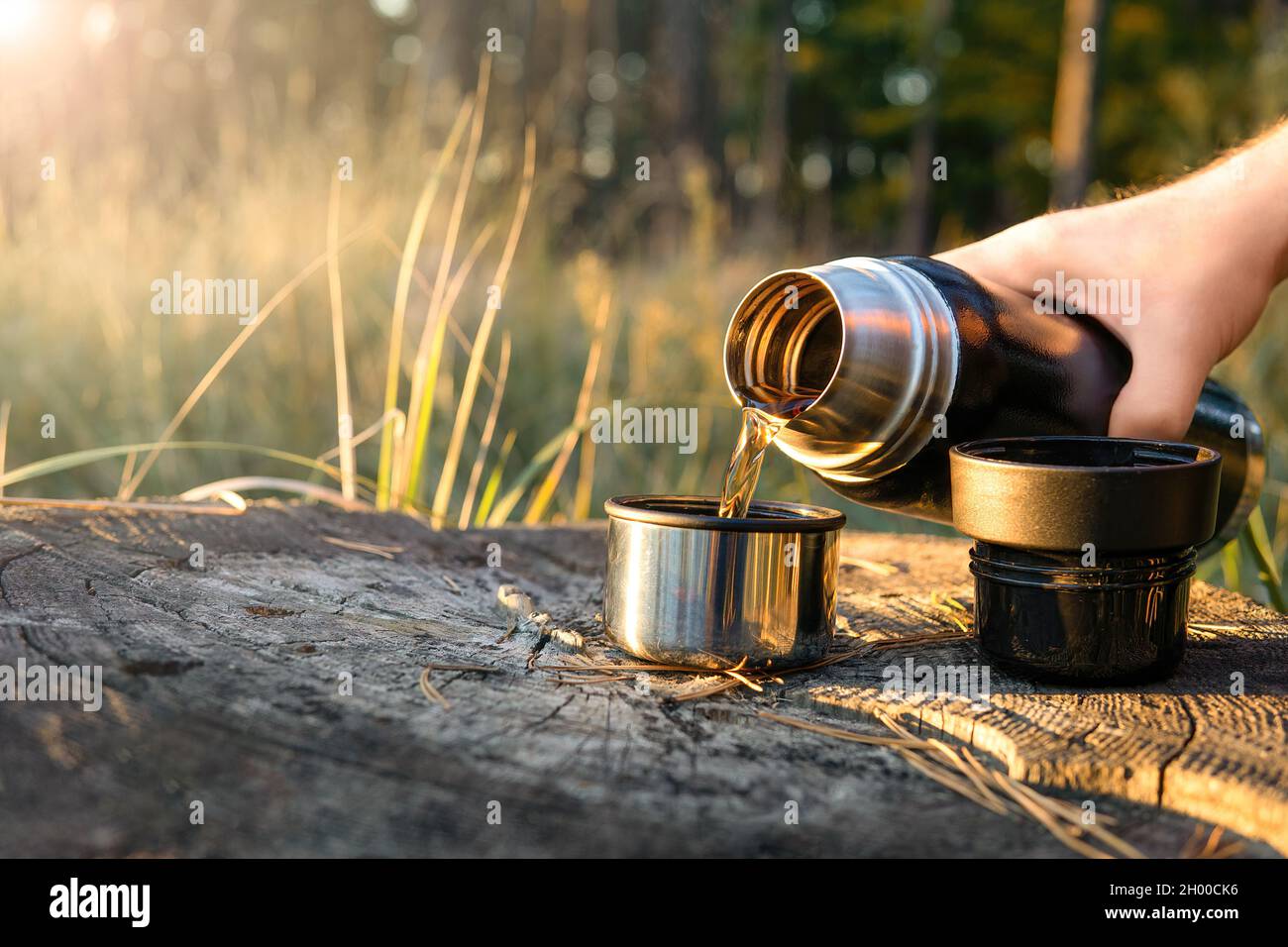 Il turista versa il tè caldo da una fiasca thermos in due tazze in piedi su un ceppo dell'albero al tramonto. Escursioni, viaggi e stile di vita attivo concetto Foto Stock
