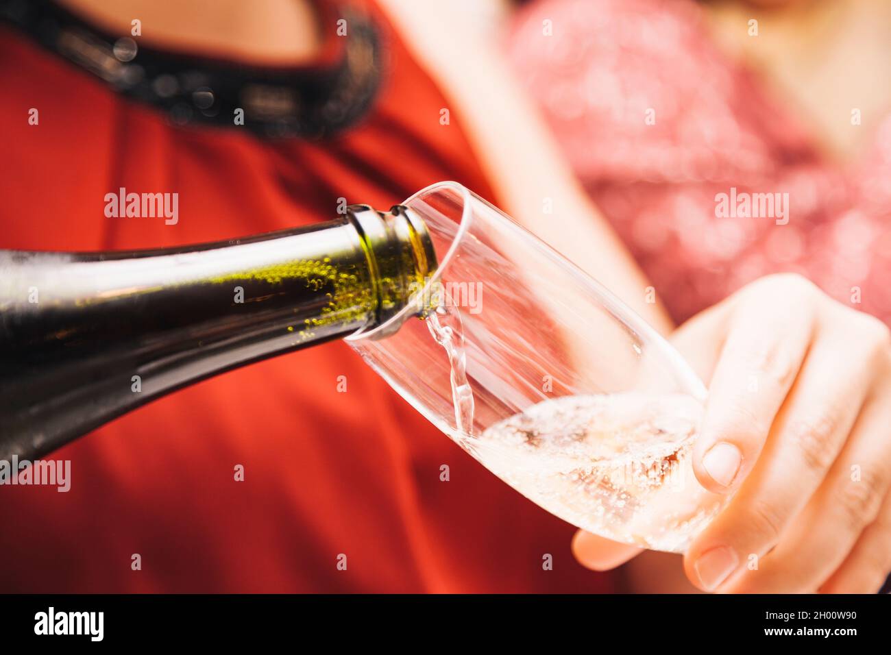 Le mani di una donna sconosciuta che indossa un vestito rosso che tiene un bicchiere mentre lo riempie di Cava da una bottiglia di vetro verde. Foto Stock