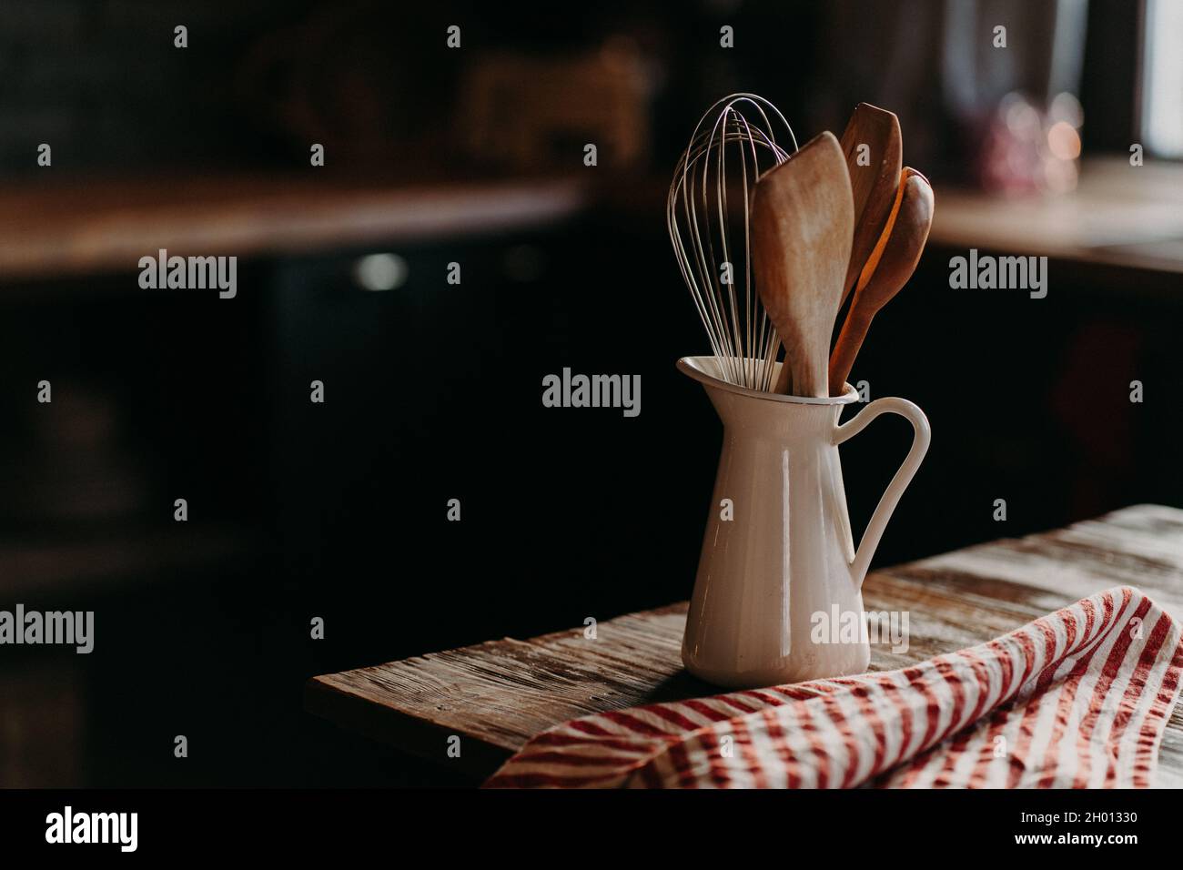 Accessori da cucina su tavolo in legno. Utensili in vaso di ceramica bianca su sfondo scuro. Stile rustico. Stoviglie per preparare il pasto. Cucchiai di legno Foto Stock