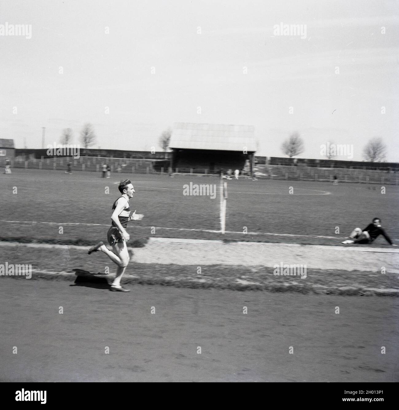 Anni '50, storico, in un campo sportivo, un giovane atleta maschile che corre fuori su una pista di cinder o sterrato, andando passare il sandpit del salto lungo, Inghilterra, Regno Unito. Foto Stock