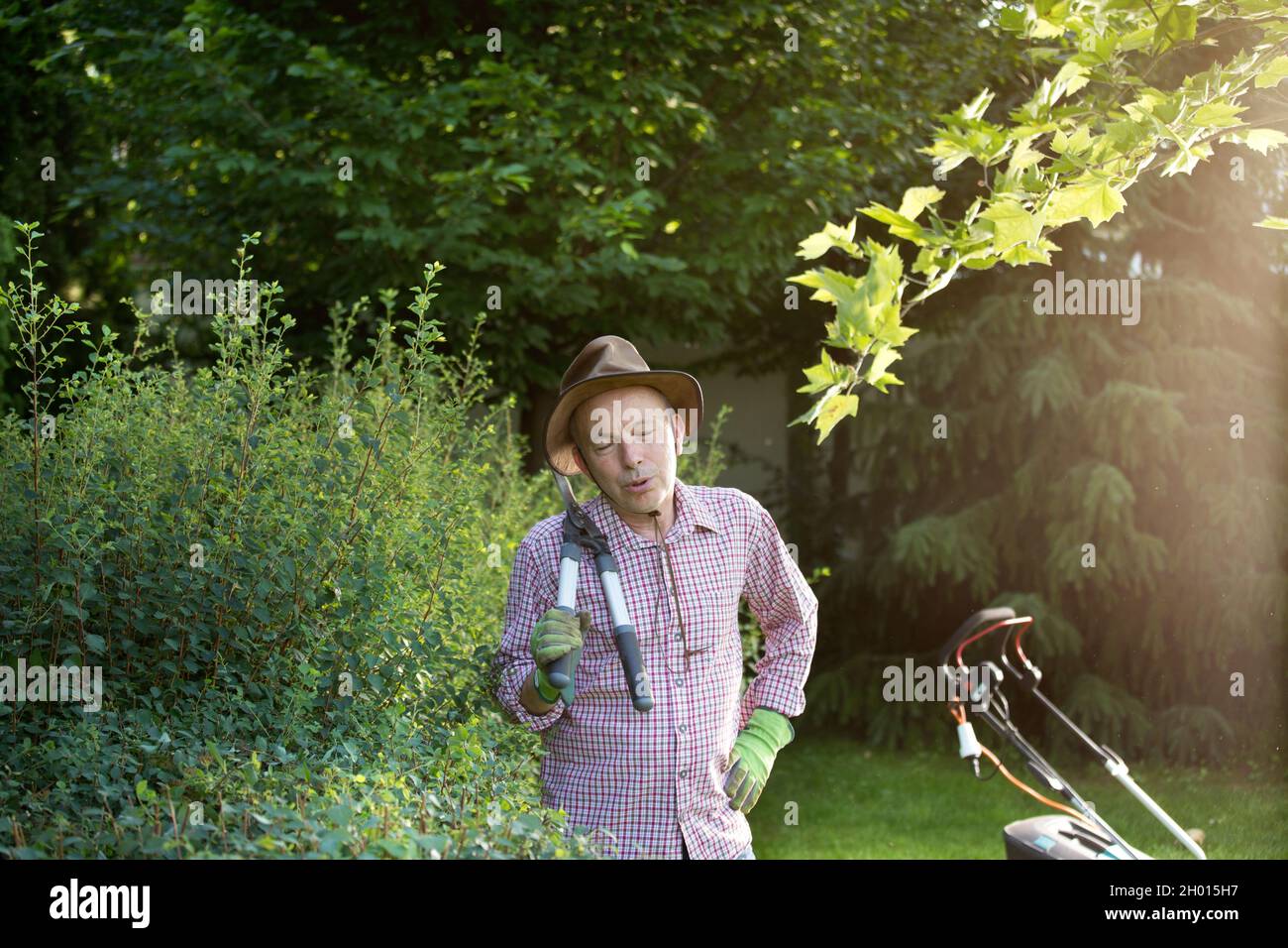 Ritratto di uomo stanco con forbici da giardinaggio in piedi accanto alla siepe con rasaerba sullo sfondo. Prendersi cura di piante Foto Stock