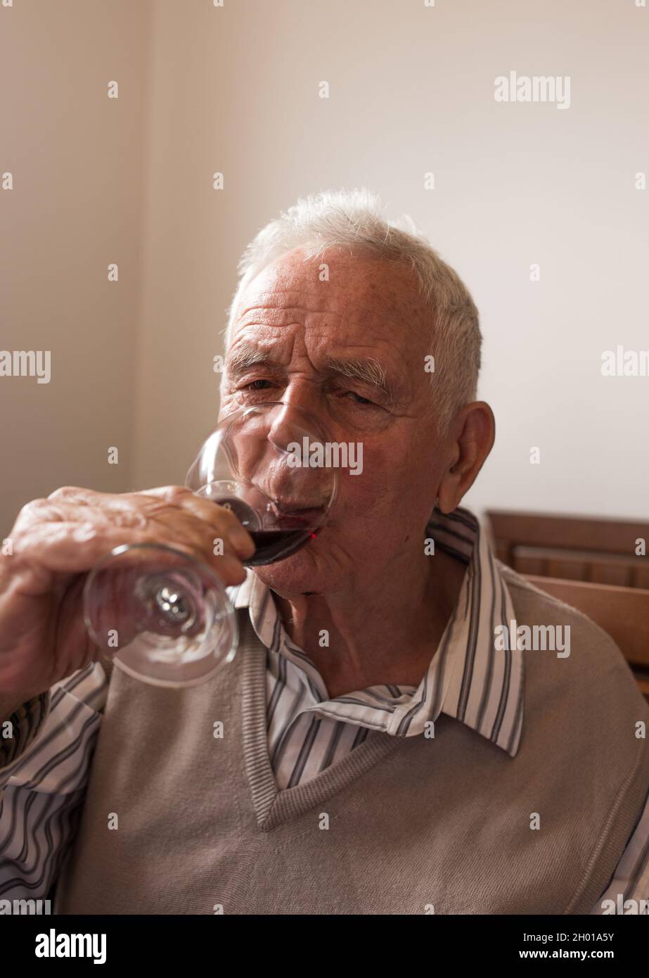 Uomo anziano in camicia e gilet bevendo vino rosso dal bicchiere a casa Foto Stock