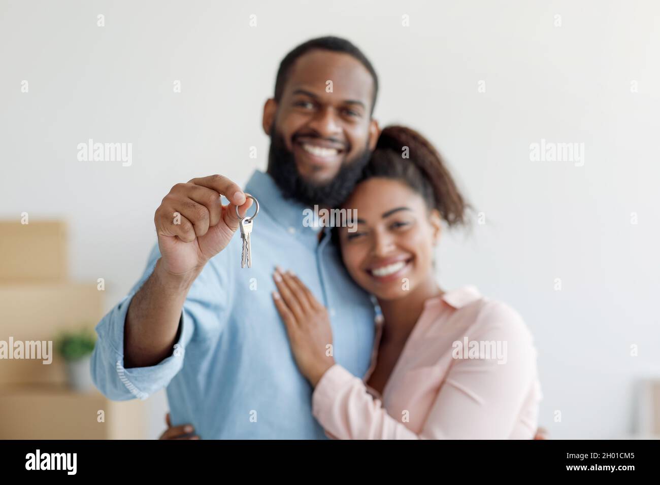 Il giovane e allegro marito afroamericano abbraccia la moglie e mostra le chiavi in camera con scatole di cartone. Coppia positiva godersi l'acquisto di nuova casa e il loro proprio hou Foto Stock