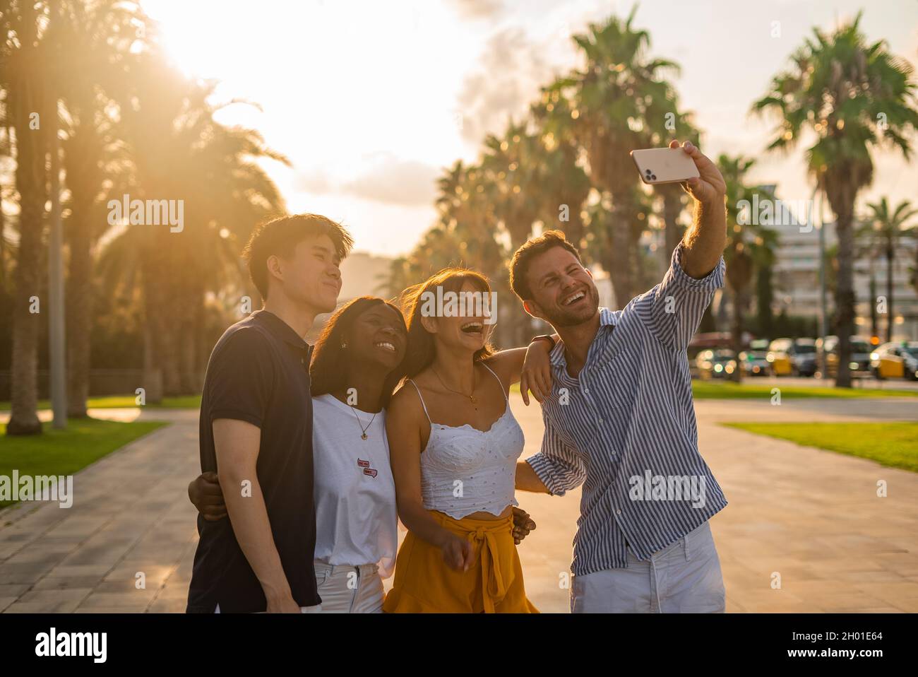 Gruppo di giovani studenti multirazziali che si divertono e prendono selfie su smartphone durante l'incontro su strada urbana con palme verdi nella soleggiata serata estiva Foto Stock