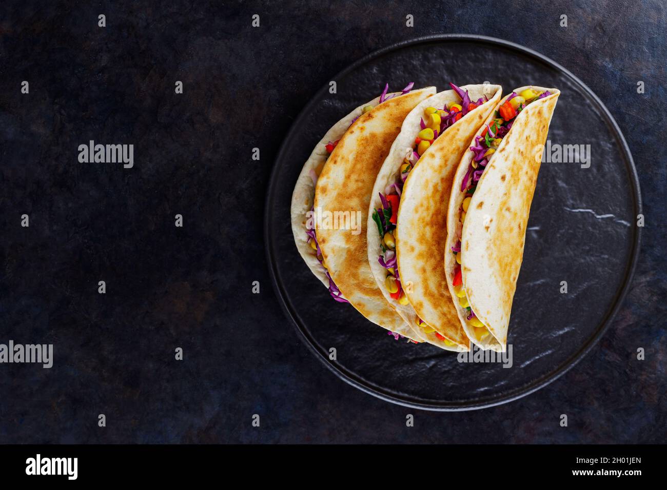 Tacos vegetariani con mais dolce, cavolo viola e pomodori su un piatto nero. Tacos con verdure e salsa guacamole su sfondo scuro. Copia spa Foto Stock