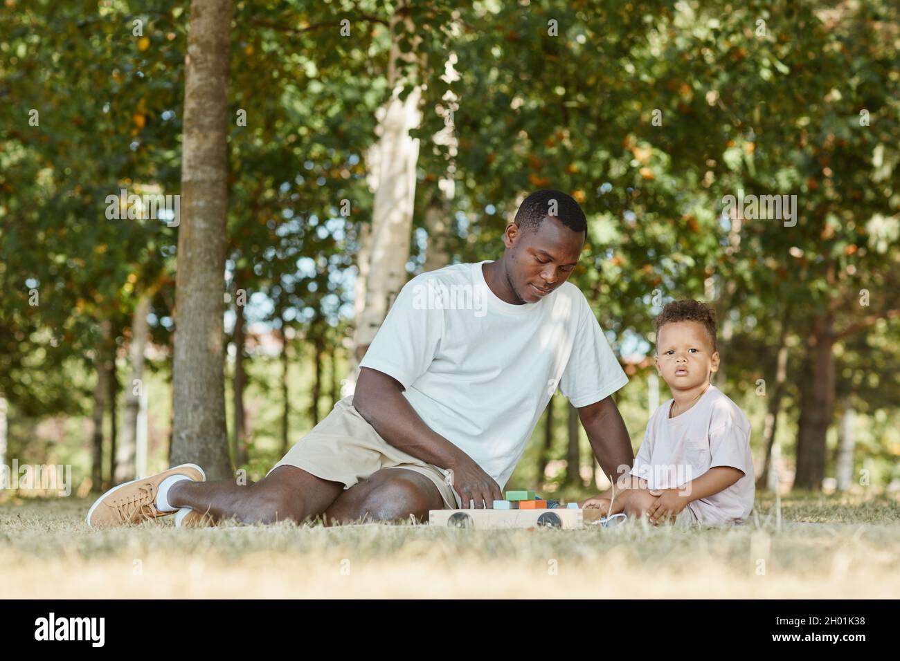 Ritratto del giovane padre afroamericano che gioca con il figlio carino nel parco mentre si siede sull'erba, spazio copia Foto Stock