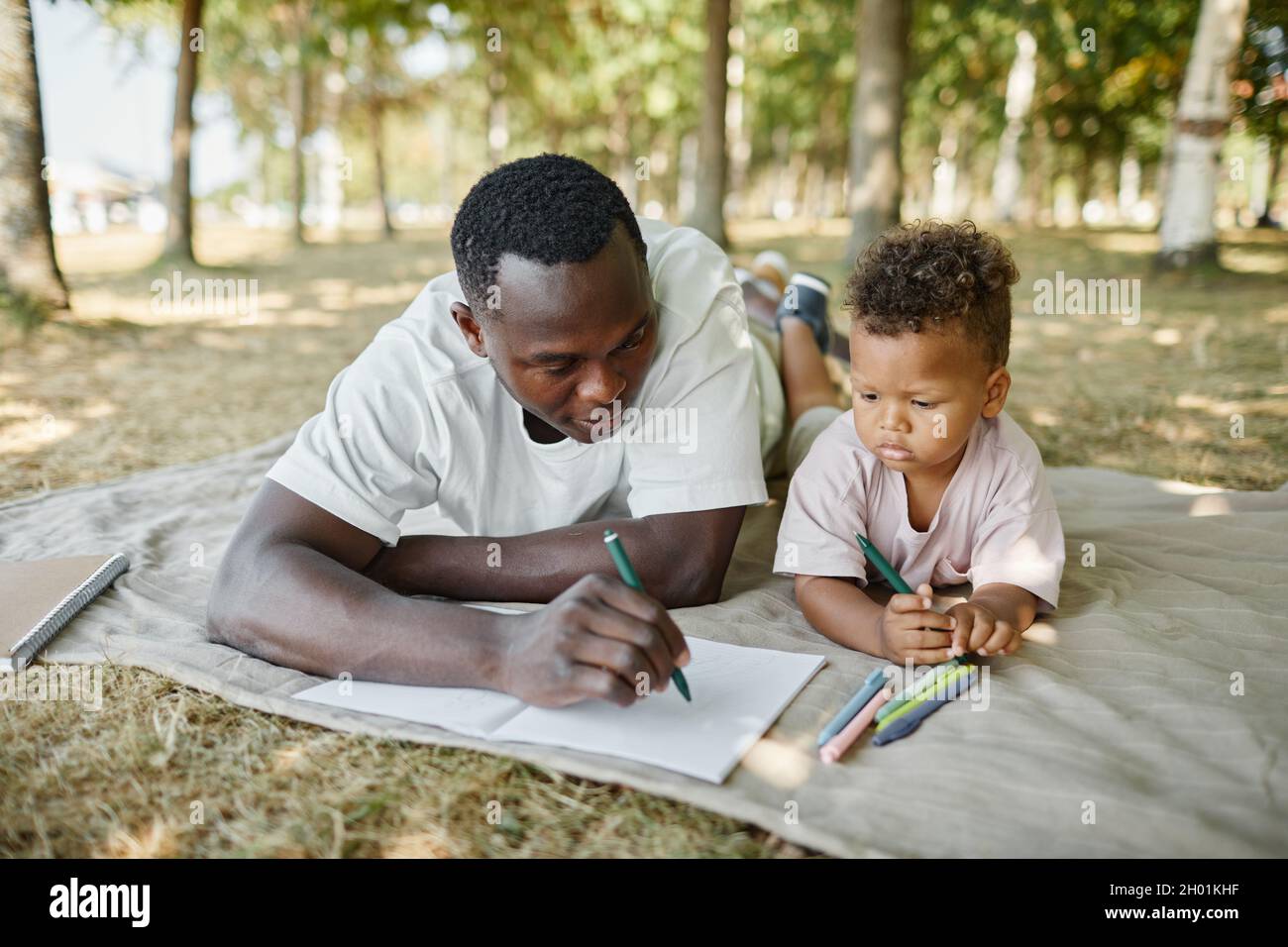 Ritratto del giovane padre afroamericano che gioca con il figlio carino nel parco e che si disegna insieme mentre si posa su erba, spazio copia Foto Stock