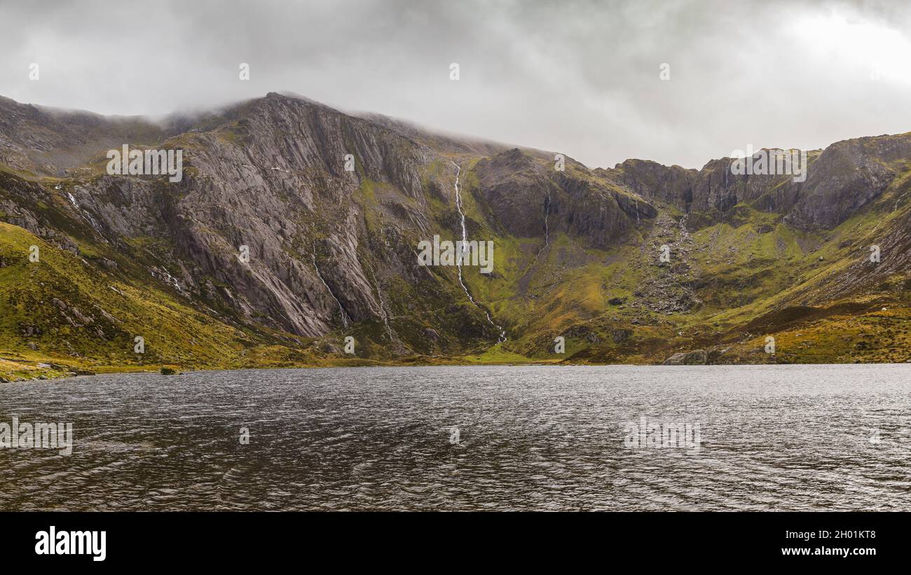 La luce del sole pierce le nuvole scure sul Lago Idwal nel Parco Nazionale di Snowdonia che illuminano il lato della montagna e i corsi d'acqua piovana che scendono. Foto Stock