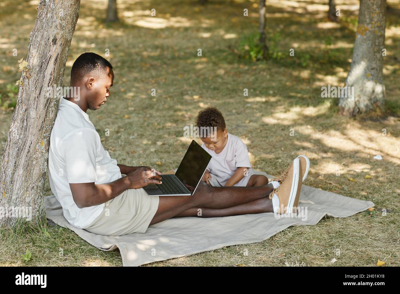 Ritratto vista laterale di padre afro-americano usando il laptop mentre gioca con il figlio carino nel parco, spazio copia Foto Stock