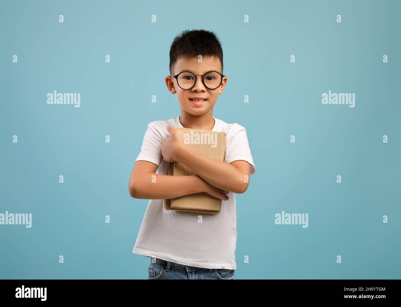 Carino ragazzo asiatico piccolo indossare occhiali che tengono stack di libri e guardando la macchina fotografica, nerdy coreano maschio bambino godendo di lettura e apprendimento, in piedi Foto Stock