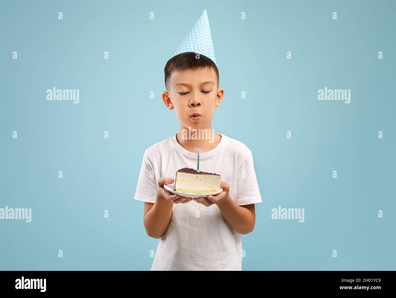 Carino ragazzo asiatico piccolo in parte cappello che soffia candela sulla torta di compleanno, bambino piccolo felice maschio che tiene pezzo di dolce B-giorno, facendo il desiderio mentre si levano in piedi Foto Stock