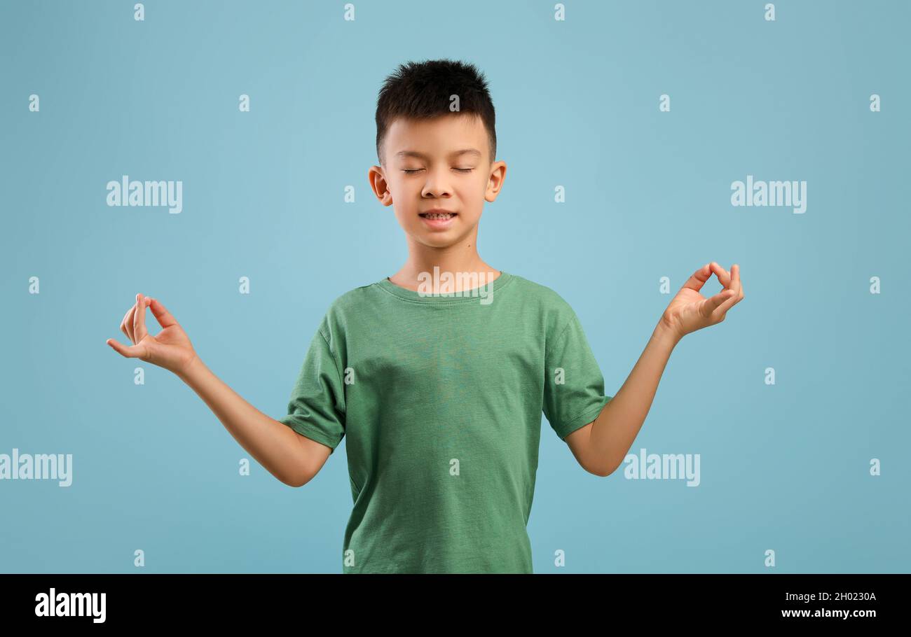 Zen. Rilassato ragazzo asiatico meditando con occhi chiusi su sfondo blu, calmo coreano maschio bambino praticare yoga, mantenendo le mani in Mudra Gesture Foto Stock