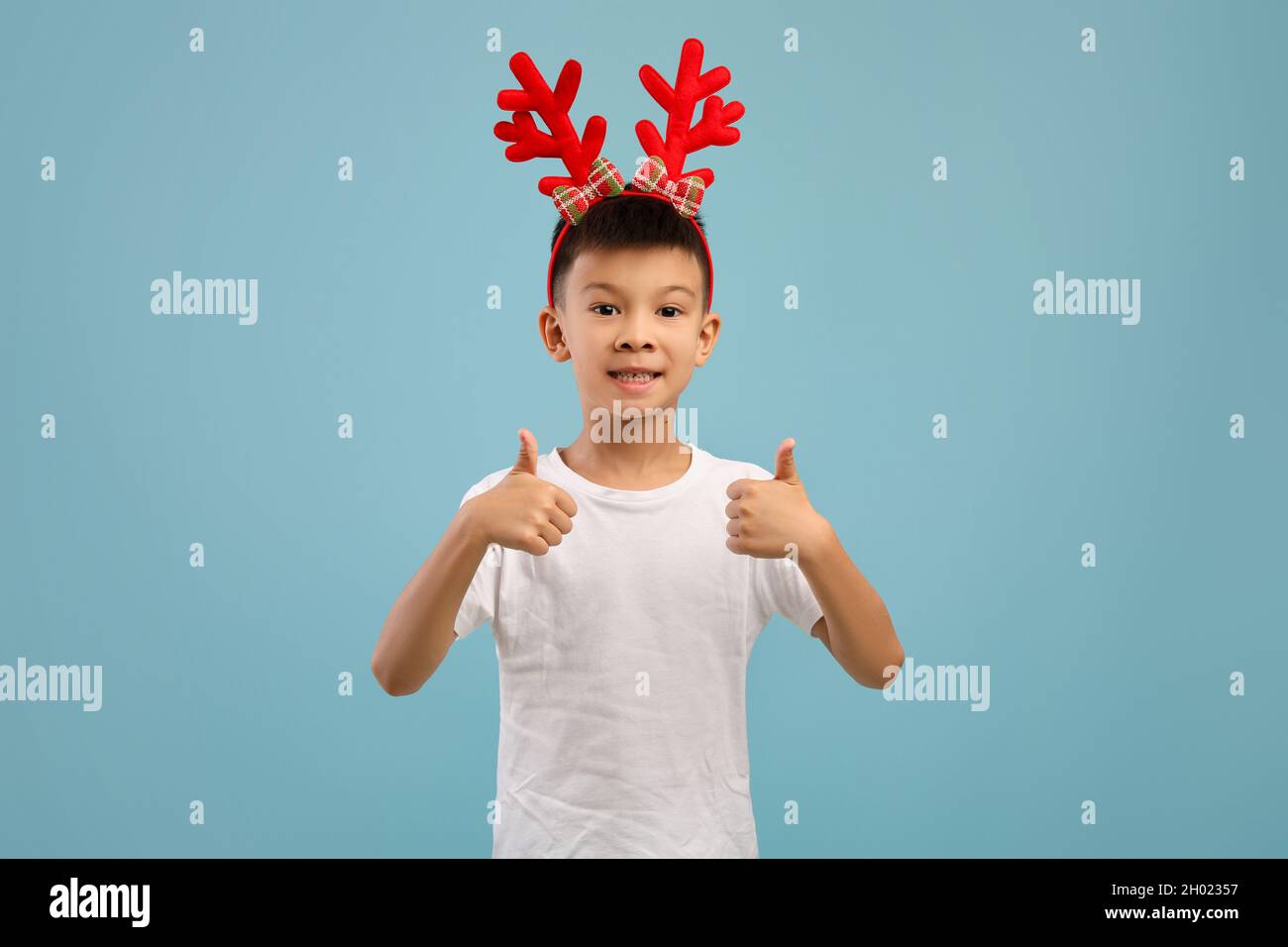 Carino ragazzo asiatico piccolo indossare Xmas Deer Horns gesturing Thumbs in su a macchina fotografica, sorridente bambino maschio di Premeen raccomandare l'offerta di festa di Natale mentre sta Foto Stock