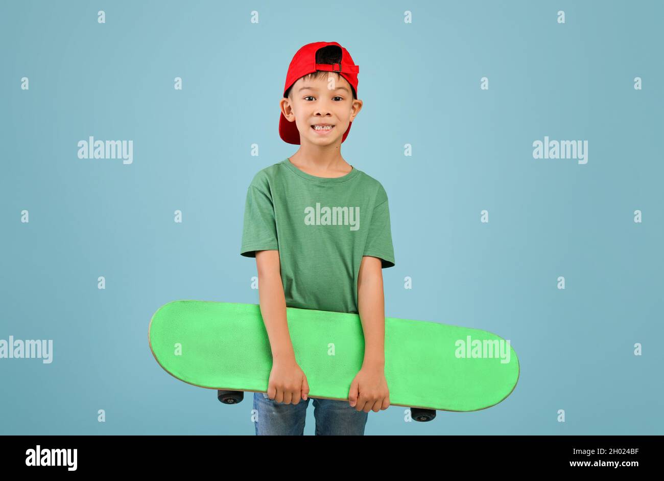 Allegro eccitato ragazzo asiatico piccolo in rosso cappello tenendo skateboard e guardando la macchina fotografica, felice sorridente coreano bambino maschio in abiti casual in posa isolat Foto Stock