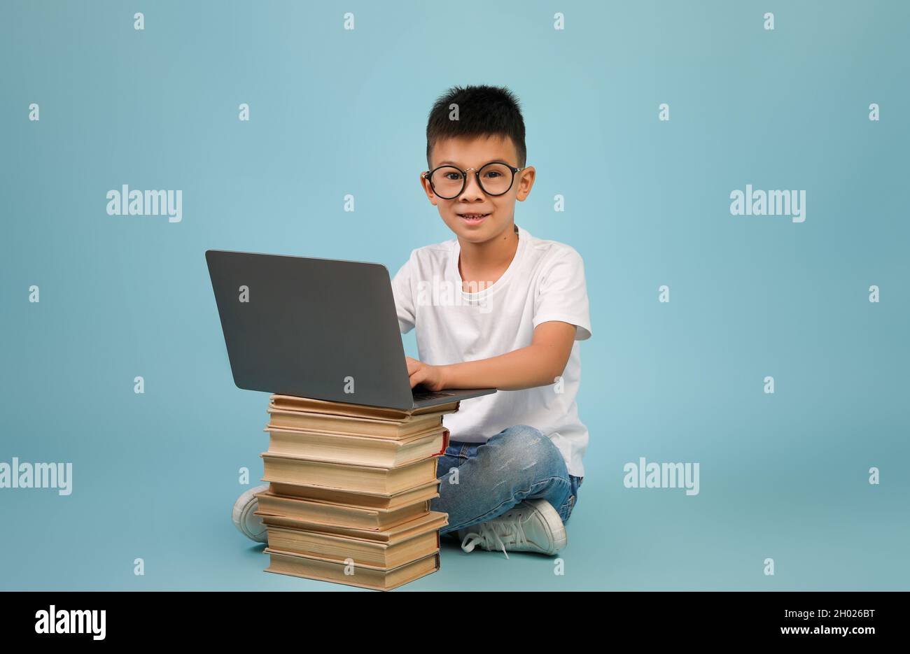 Piccolo ragazzo asiatico che indossa occhiali seduti con laptop e pila di libri e guardando la macchina fotografica, coreano maschio bambino che gode di formazione a distanza, Posin Foto Stock