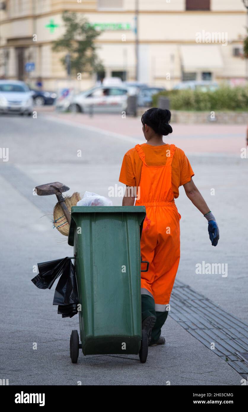 Vista posteriore di donna spazzatura lavoratore in arancione uniforme tirando cestino con altre attrezzature Foto Stock