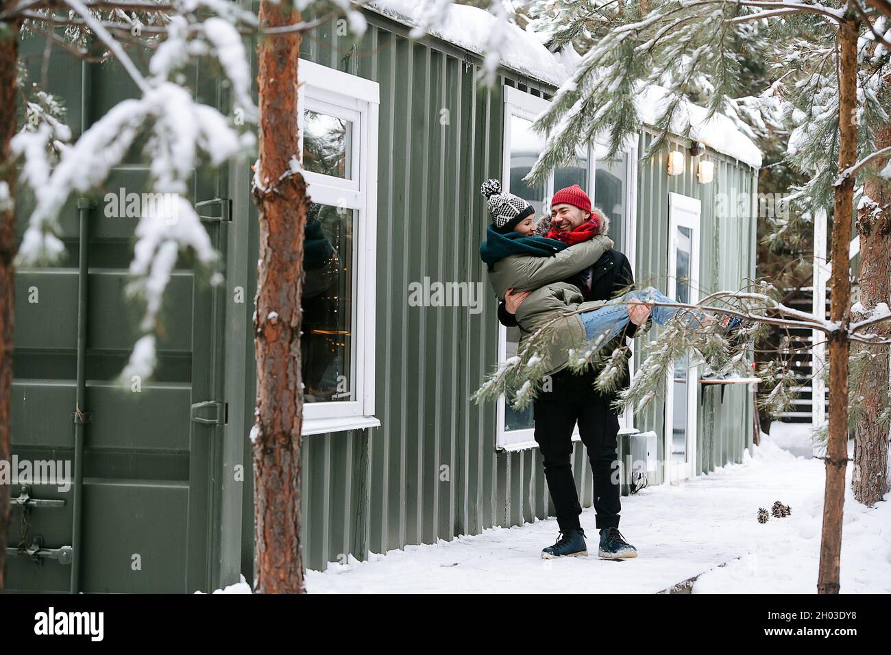 Coppia amorevole all'aperto in inverno. L'uomo porta la donna nelle sue mani. Entrambi indossano cappelli, giacche e sciarpe. Di fronte ad una casa con corrugato s Foto Stock