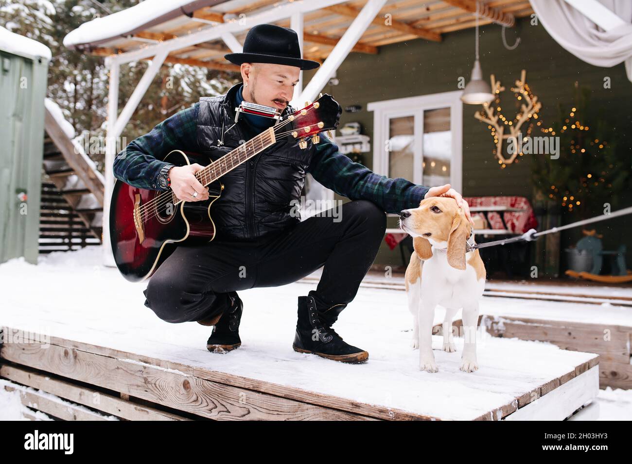 Musicista stagionato in un cappello e giacca di pelle che pattina un cane sulla testa. Sta accovacciandosi davanti alla casa su un pavimento innevato della tavola. Foto Stock