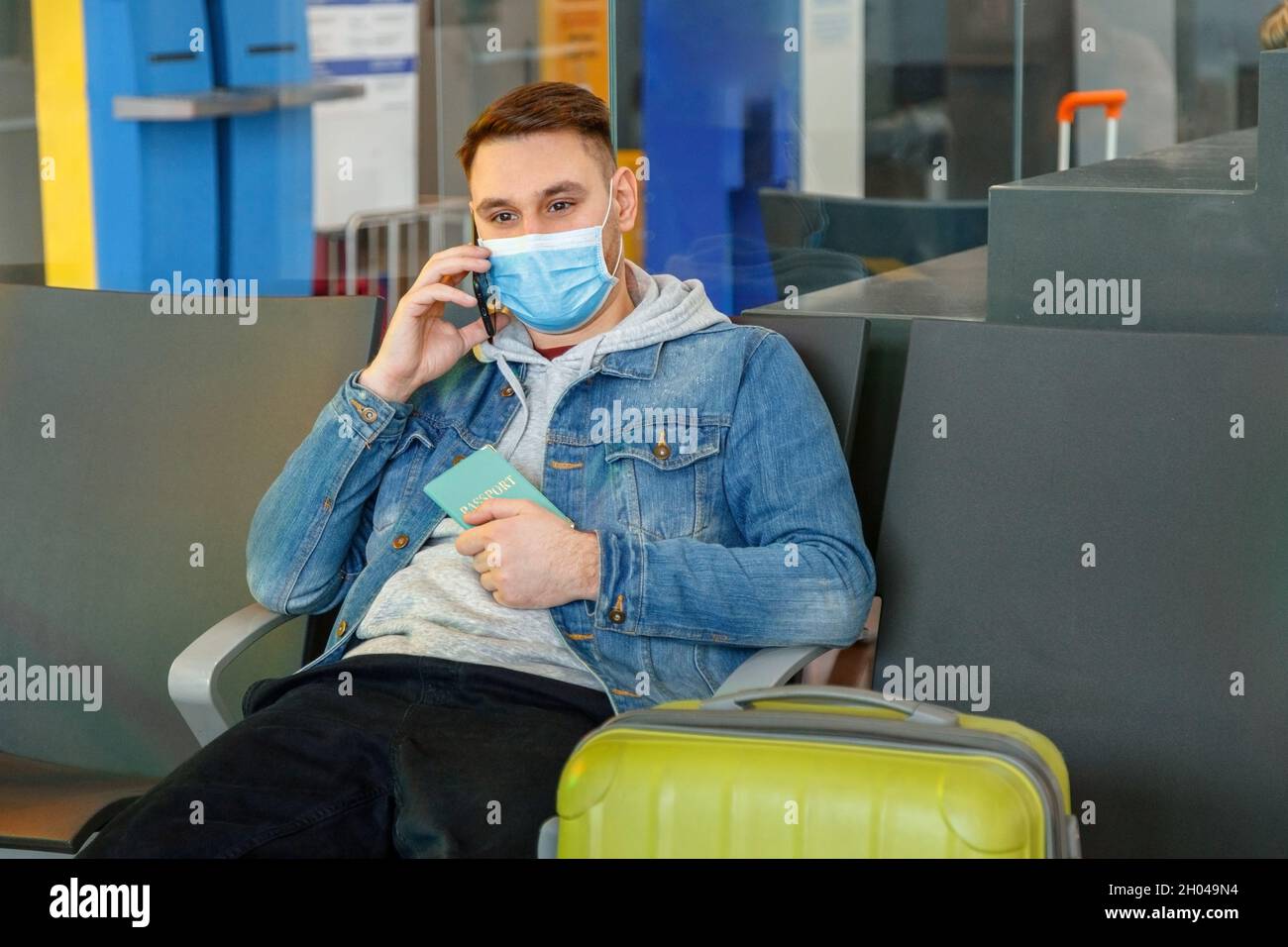 Giovane uomo attraente in maschera medica parlare con smartphone nella lounge dell'aeroporto con valigia che tiene il passaporto in mano prima di viaggiare Foto Stock
