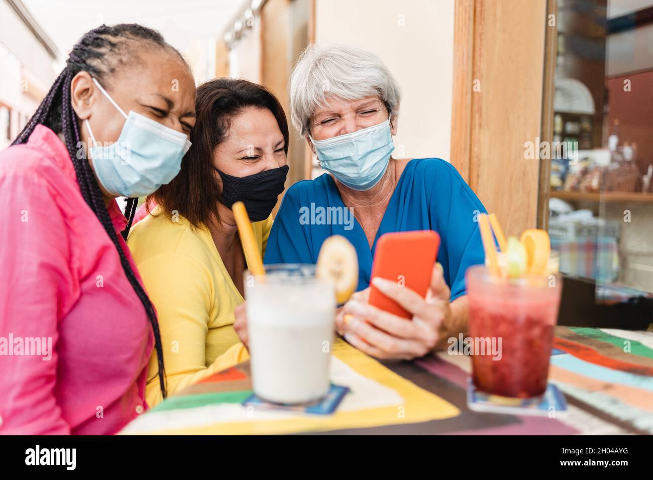 Amici anziani multirazziali che usano il telefono cellulare al ristorante brunch che indossa maschere di sicurezza - persone di generazione anziana, concetto di tecnologia - Focus su RIG Foto Stock