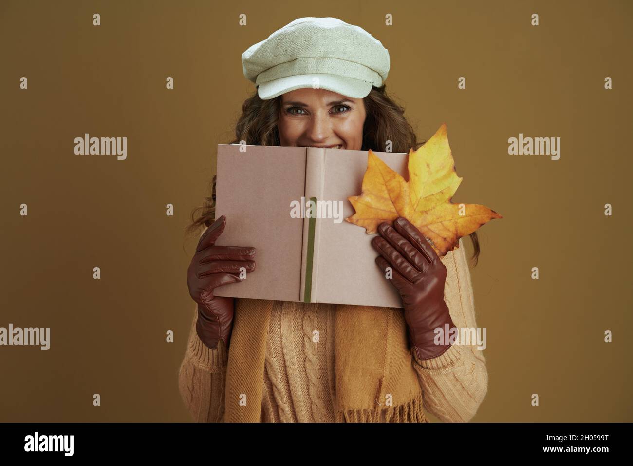 Ciao settembre. Felice elegante donna di mezza età in sciarpa con foglia di acero giallo autunno, libro e guanti in pelle su sfondo marrone. Foto Stock