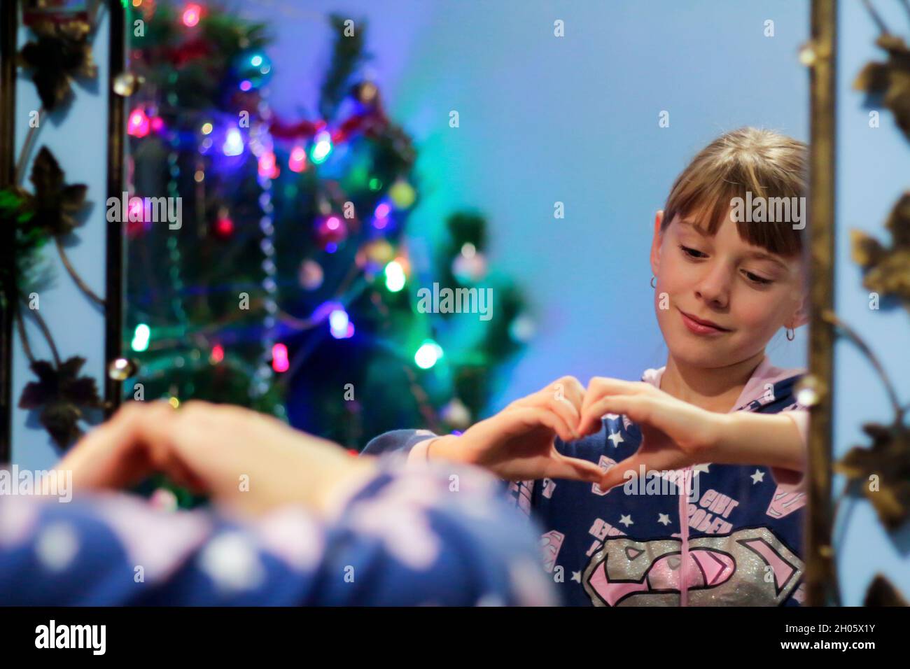 Defocus bionda ragazza caucasica 10 anni in pigiama mostra il cuore del gesto su riflessione specchio. Verticale sullo specchio. Riflessione. Anno nuovo. Bambino Foto Stock
