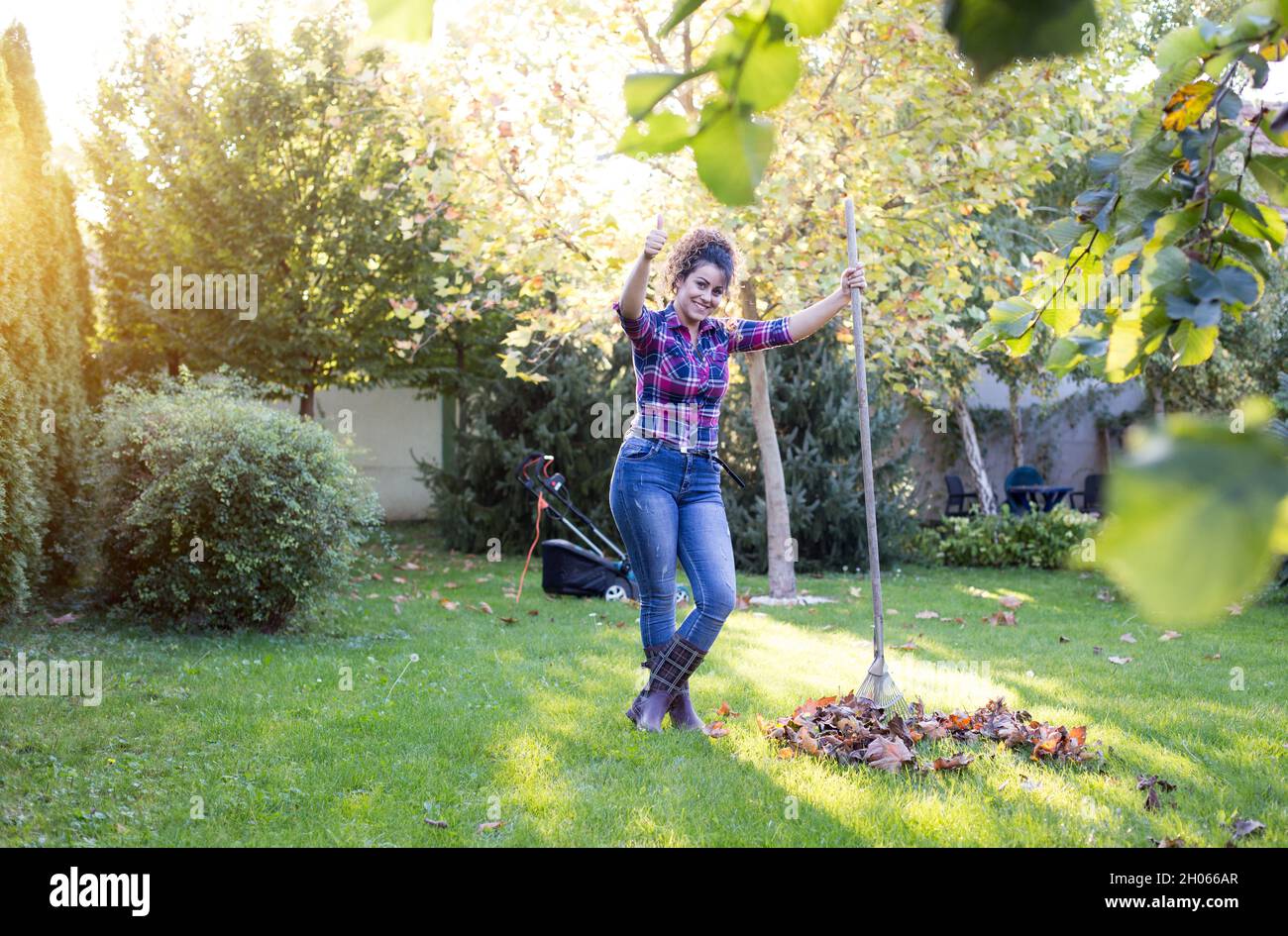 Donna graziosa che rastrellava foglie in giardino e mostrando pollice in su Foto Stock