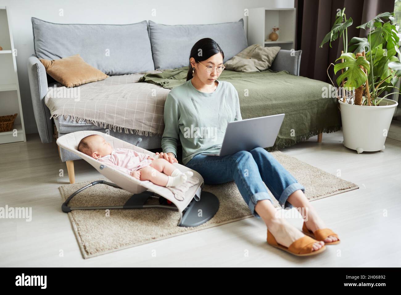 Giovane donna asiatica seduta sul pavimento nella stanza che lavora in linea sul laptop e si prende cura di sua figlia piccola Foto Stock