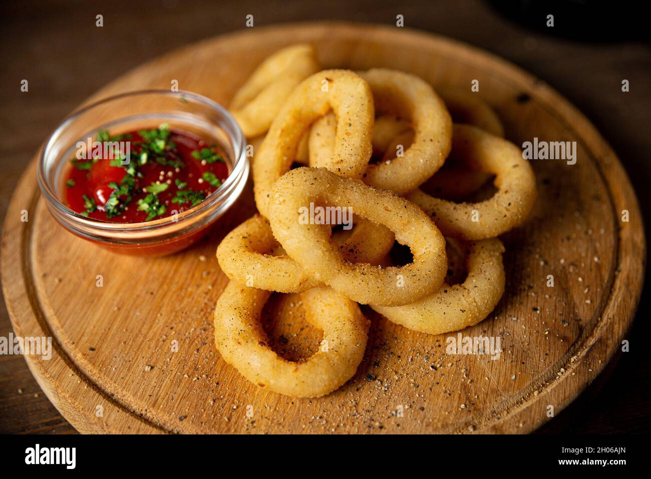 Anelli di cipolla con ketchup su un tagliere di legno. Cibo da bar. Un servizio a persona. Cospargere con pepe macinato. Un piccolo mazzo di anelli di cipolla. Foto Stock