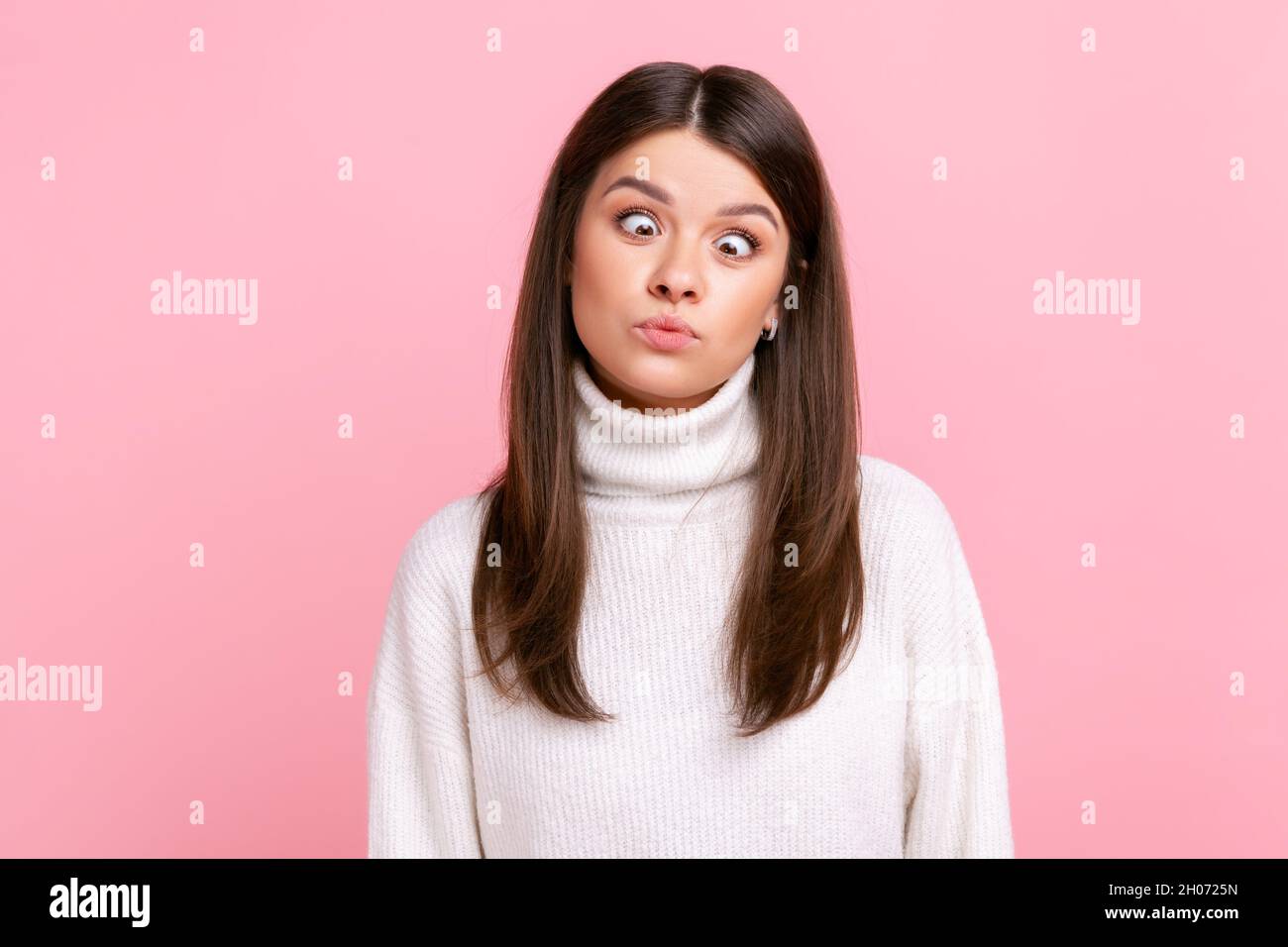 Donna idiota che fa il viso sciocco e umoroso con gli occhi incrociati, mostrando comico silly espressione senza cervello, indossando un pullover bianco casual stile. Studio interno girato isolato su sfondo rosa. Foto Stock