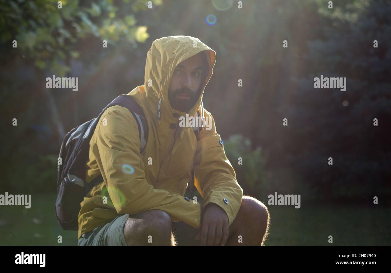 Giovane uomo attraente con barba seduta su roccia nella foresta con impermeabile e cappuccio sulla testa Foto Stock