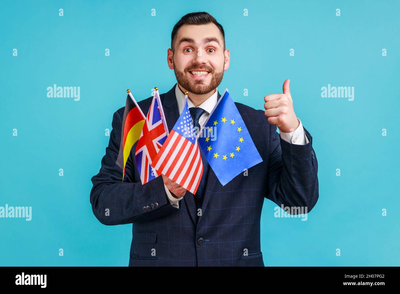 Ritratto di uomo d'affari felice portatore di vestito di stile ufficiale che tiene bandiere di Germania, Stati Uniti, Gran Bretagna ed Unione europea, mostrando pollice in su. Studio interno girato isolato su sfondo blu. Foto Stock