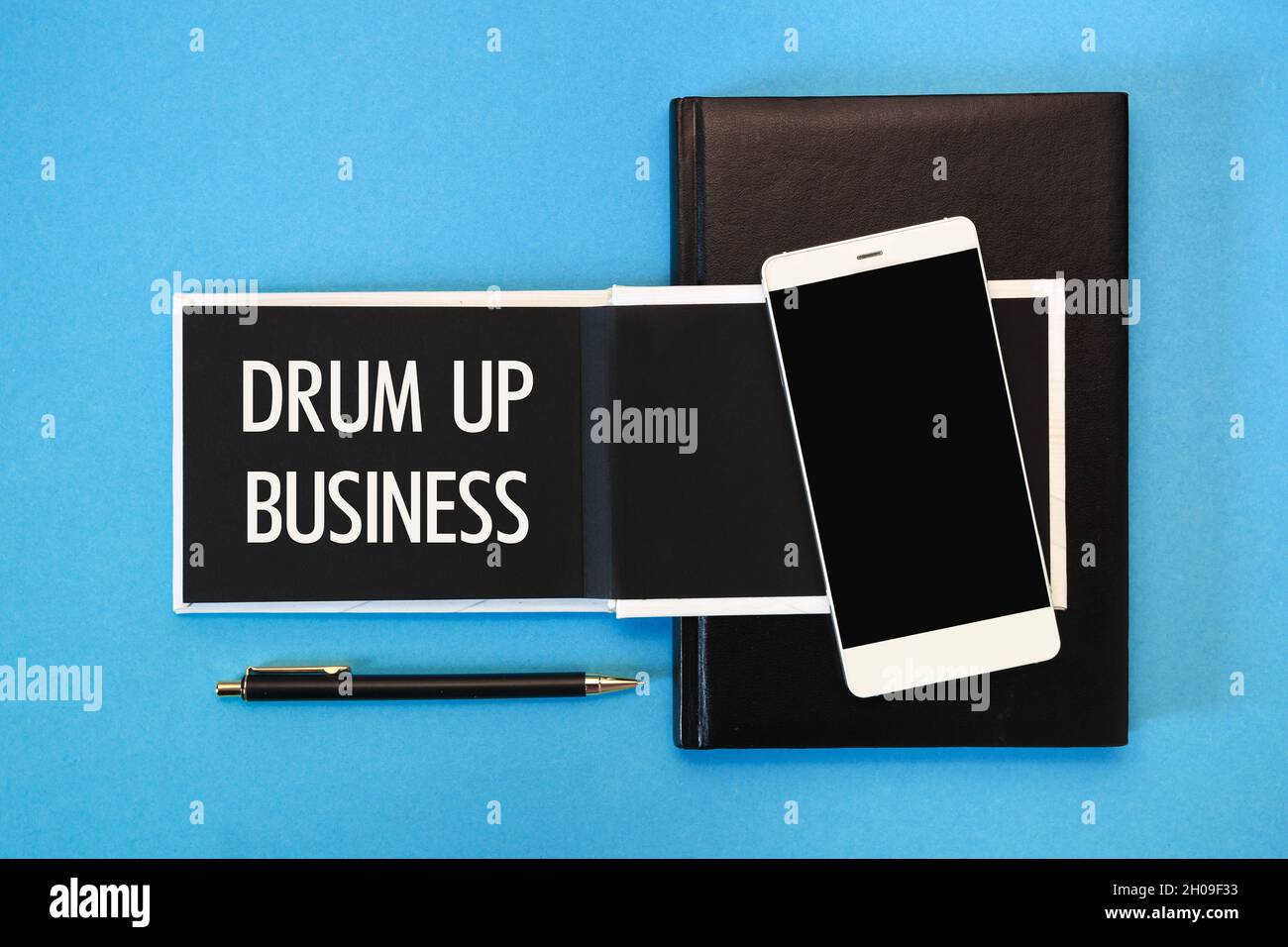 Un notebook bianco con pagine nere, uno smartphone e una penna su sfondo blu. L'iscrizione Drum Up Business sul blocco note. Foto Stock