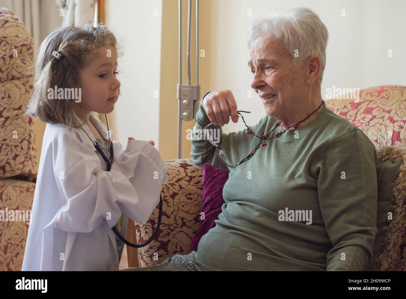 Una bambina in un cappotto medico esamina la nonna con uno stetoscopio. Giochi per bambini. Focu Girl selettivo Foto Stock